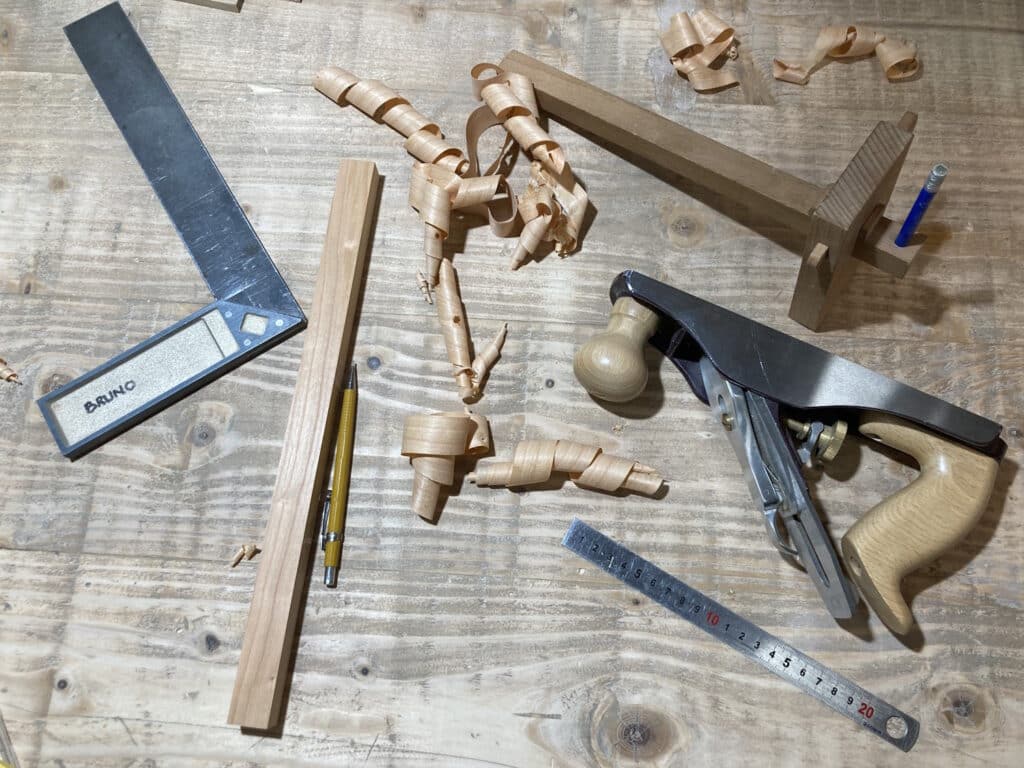 Cette image montre une vue d'un dessus d'établi de menuisier sur lequel se trouve quelques outils et instruments typiques du travail du bois: rabot, trusquin, équerre règlet et crayon. Quelques copeaux bouclés sont éparpillés entre les outils.