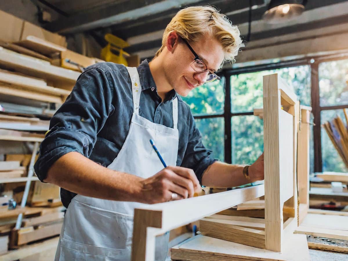 Cette image montre un jeune menuisier au travail dans son atelier. Il porte des lunettes et un tablier de travail. Il trace au crayon sur un cadre en bois