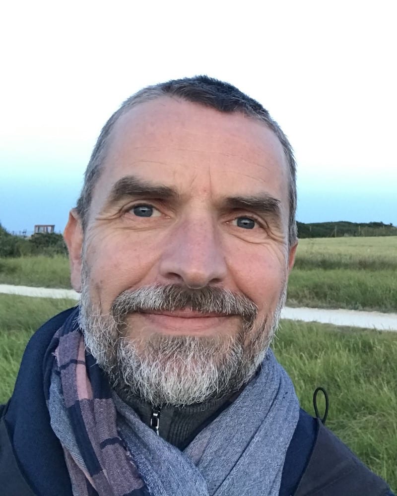Cette image est un portrait de Bruno Peytavin. C’est une photo en extérieur avec un ciel dégagé et de l’herbe verte en arrière plan. On devine qu’il fait frais à l’habillement: manteau et écharpe.