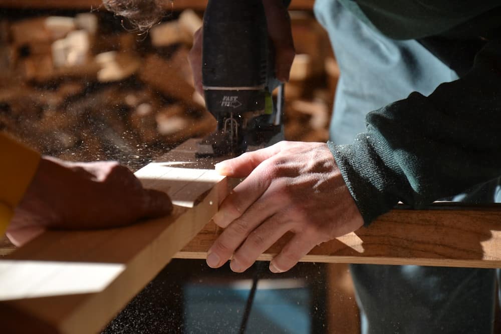 Cette image montre des mains en gros plan en train de travailler de concert. On devine qu’il y a 2 personnes dont chacune une main. L’une coupe une planche de bois avec une scie sauteuse. L’autre semble aider en tenant fermement la pièce à couper. De la sciure vole dans la lumière oblique.